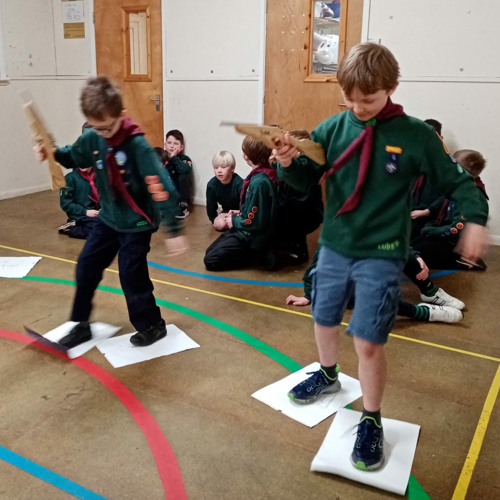 Two Cub Scouts race by sliding their feet on sheets of paper while carrying elastic-band shooters