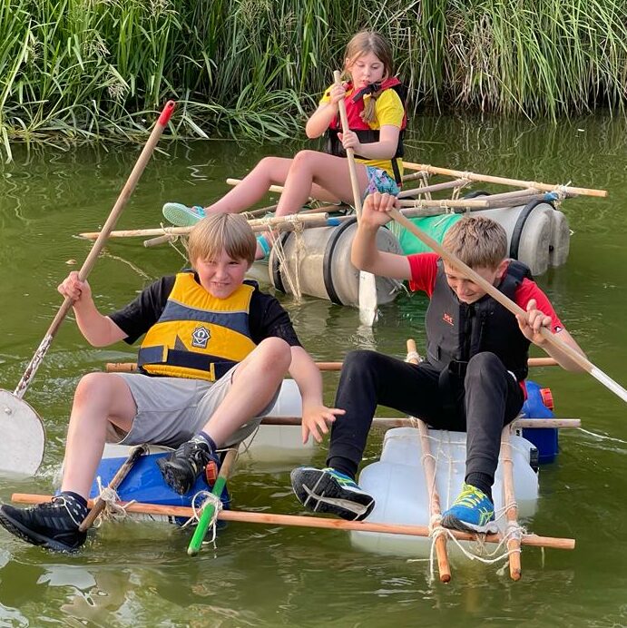 Two Scouts sitting on a home-made raft struggling to stay upright, being watched by another Scout sitting calmly on a second raft